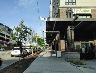 A city sidewalk beside a brick apartment building with a sandwich board sign reading Phnom Penh Noodle House, Open.