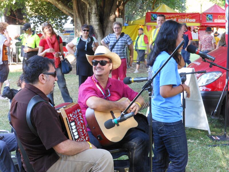 Musicians playing accordion and guitar outdoors beside a singer at a microphone, with a crowd under trees behind.