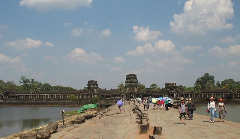 A long stone causeway between two pools leads to Angkor Wat's low entrance gallery and towers, with visitors under umbrellas and hats on the path.