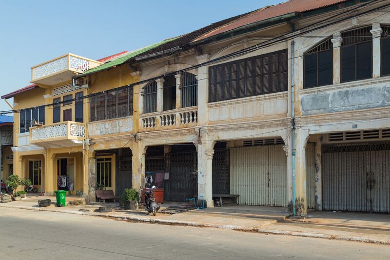 A row of weathered two-story shophouses in pale yellow and cream lines a quiet paved street, with arched arcades and pillars at street level, balconies and tall shuttered windows above, and a tiled roof. Most shopfronts are closed with folding metal gates; potted plants, a bench, a green bin and a parked motorbike stand on the sidewalk under a clear sky.