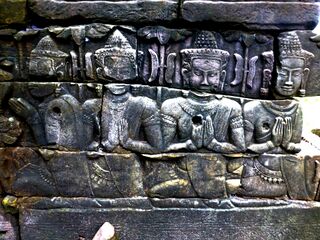 A dark sandstone relief of a row of seated figures with tall headdresses and raised hands.