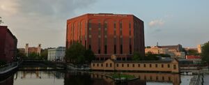 A tall red-brick college building beside a canal, with other mill buildings in the distance.