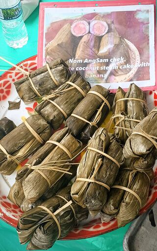 A pile of banana-leaf parcels tied with string on a red tray, a handwritten sign behind.