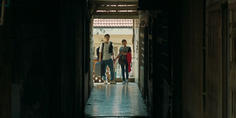 Two young people standing at the bright end of a long, dark apartment corridor.