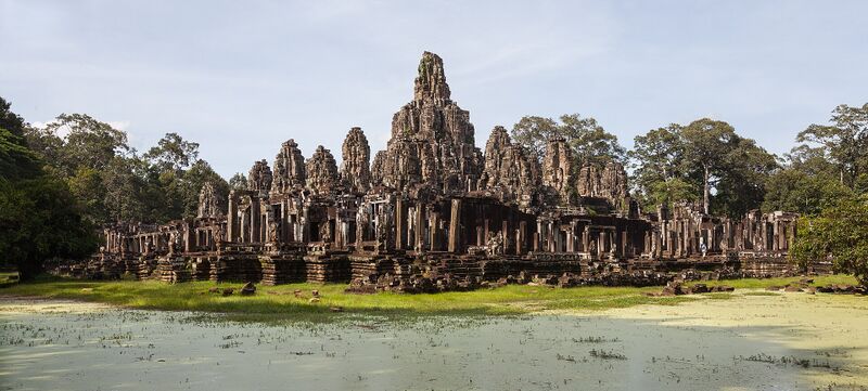 A wide view of the many towers of the Bayon rising above trees behind a green pond.