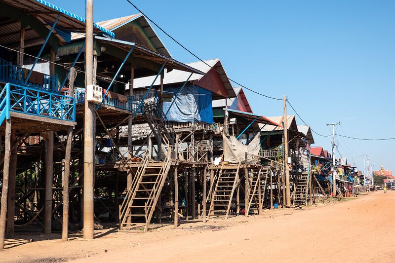A row of wooden houses raised on tall stilts, with steep ladders down to a dusty red road, under a clear blue sky.