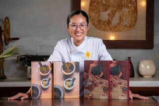 A smiling chef in a white jacket behind a counter, with four large cookbooks standing in front of her.