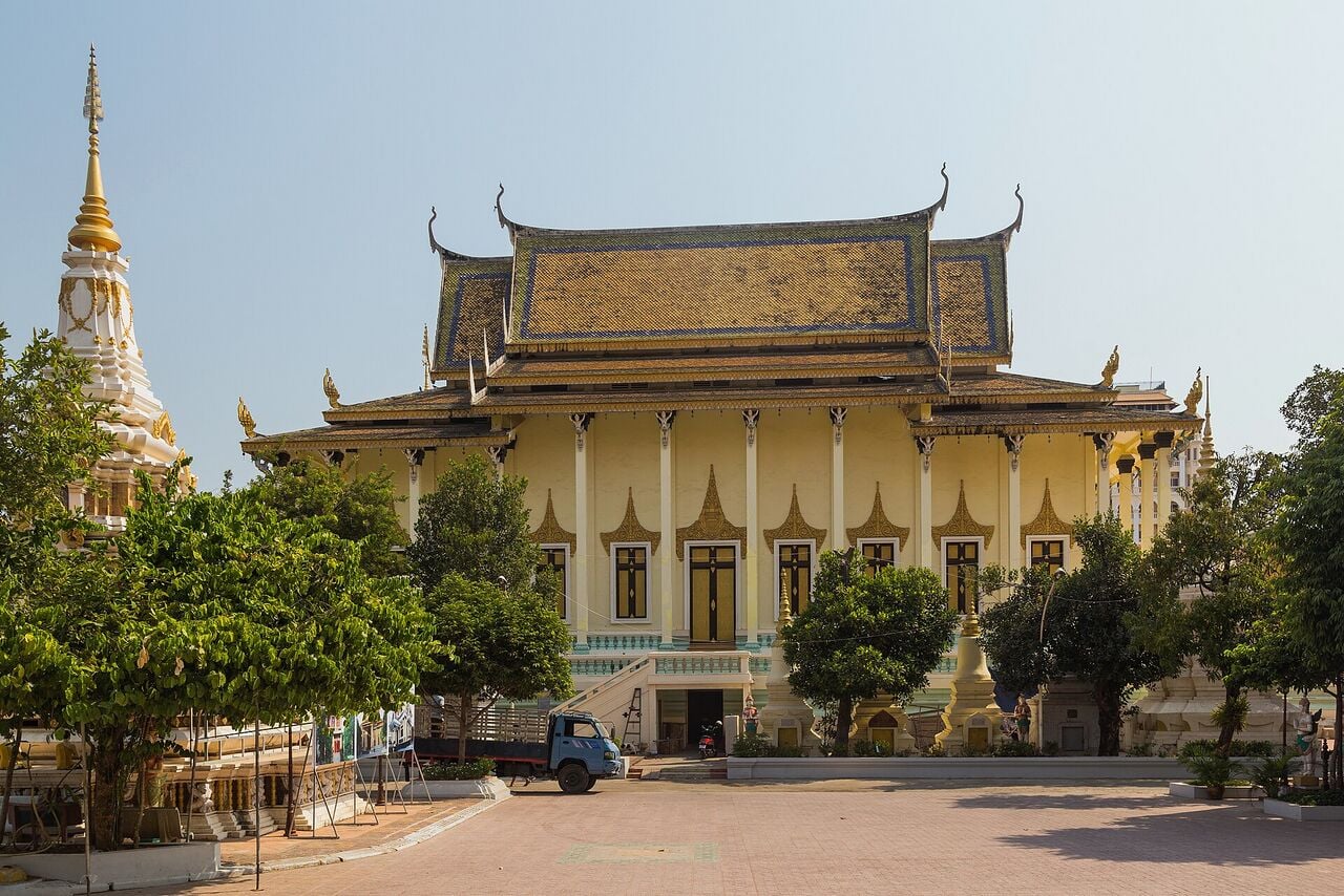 A cream-colored Buddhist prayer hall with a tiered golden roof beside a white stupa, seen across a paved courtyard