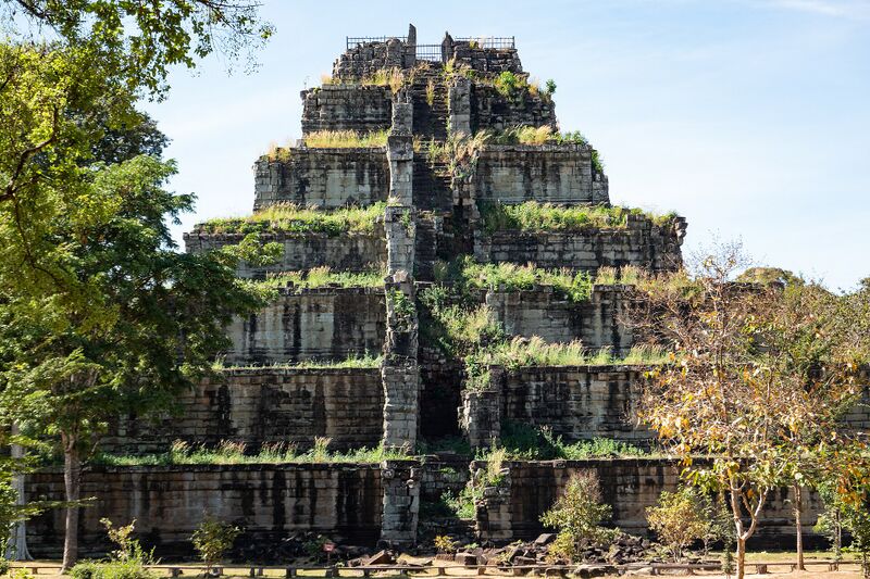 A seven-tiered stone pyramid partly covered in plants, with trees in front and a clear blue sky.