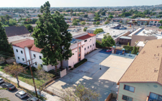 Aerial view of a city block with low buildings, a parking lot and trees.