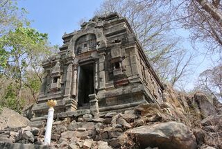 A small square stone sanctuary with a tiered roof on a rocky slope among trees.