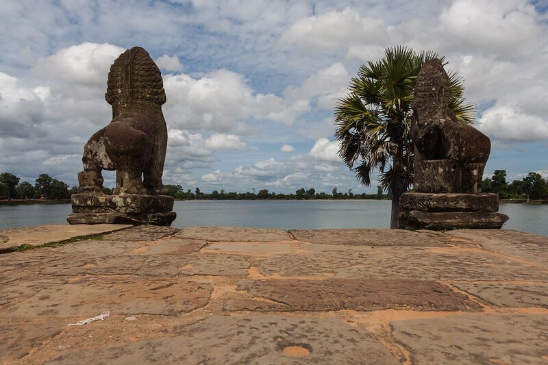 Two carved stone lions on a sandstone terrace at the edge of a wide still lake, with a palm tree and a cloudy sky.