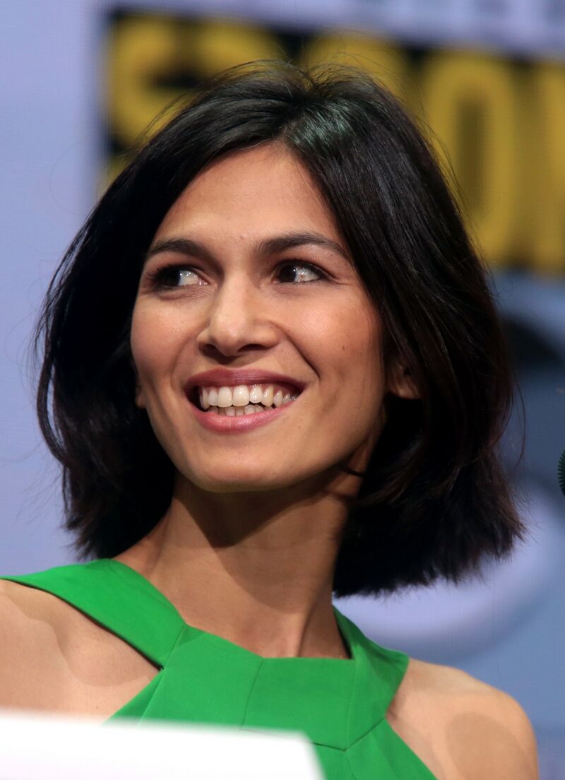 A smiling woman with shoulder-length black hair, in a green sleeveless top, looks off to the side, in front of a blurred yellow and gray backdrop.