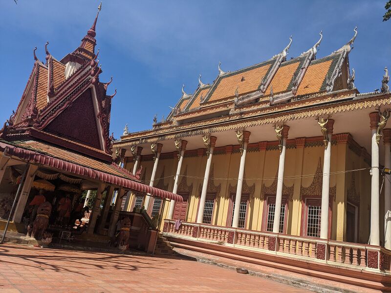 A Khmer temple hall with a tiered red roof and a row of columns, beside a tall stupa, under a blue sky.