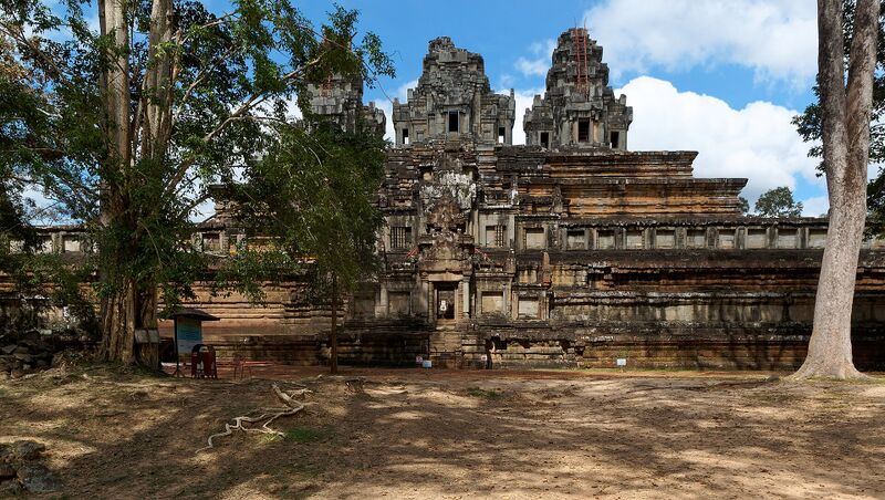 A stepped sandstone temple-mountain with five towers on its top level, seen beyond trees under a blue sky.