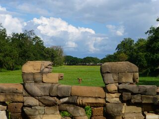Weathered stone balustrade blocks of a causeway beside a grassy moat under a cloudy sky.