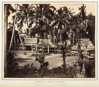 Wooden houses on stilts with thatched roofs among tall palm trees, behind a sandy open ground.