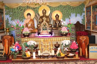 Altar with three seated Buddha images before a painted mural, decorated with flowers, candles and carved elephants.