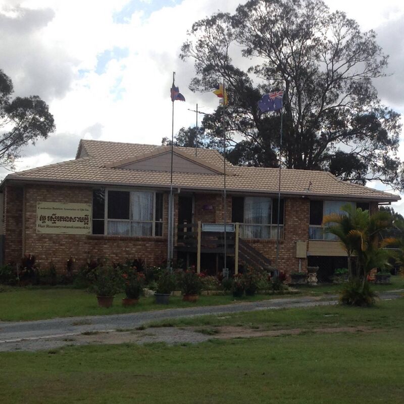 A brick building with a tiled roof and a Khmer sign, flags on poles and tall gum trees behind a lawn.