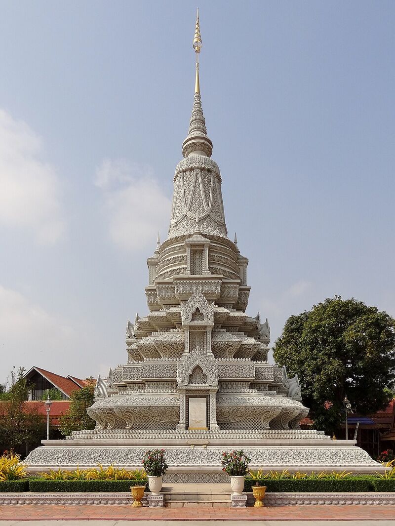 A tall white stupa with a tiered base and a slender spire, standing on a platform in a garden.