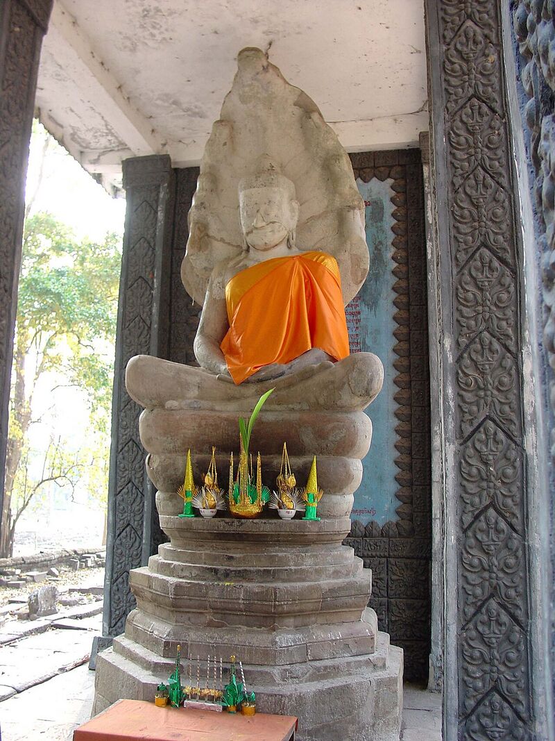 A large sandstone Buddha seated in meditation on the coils of a naga whose hoods rise behind his head, draped in an orange cloth, on a stepped stone pedestal with small offerings.