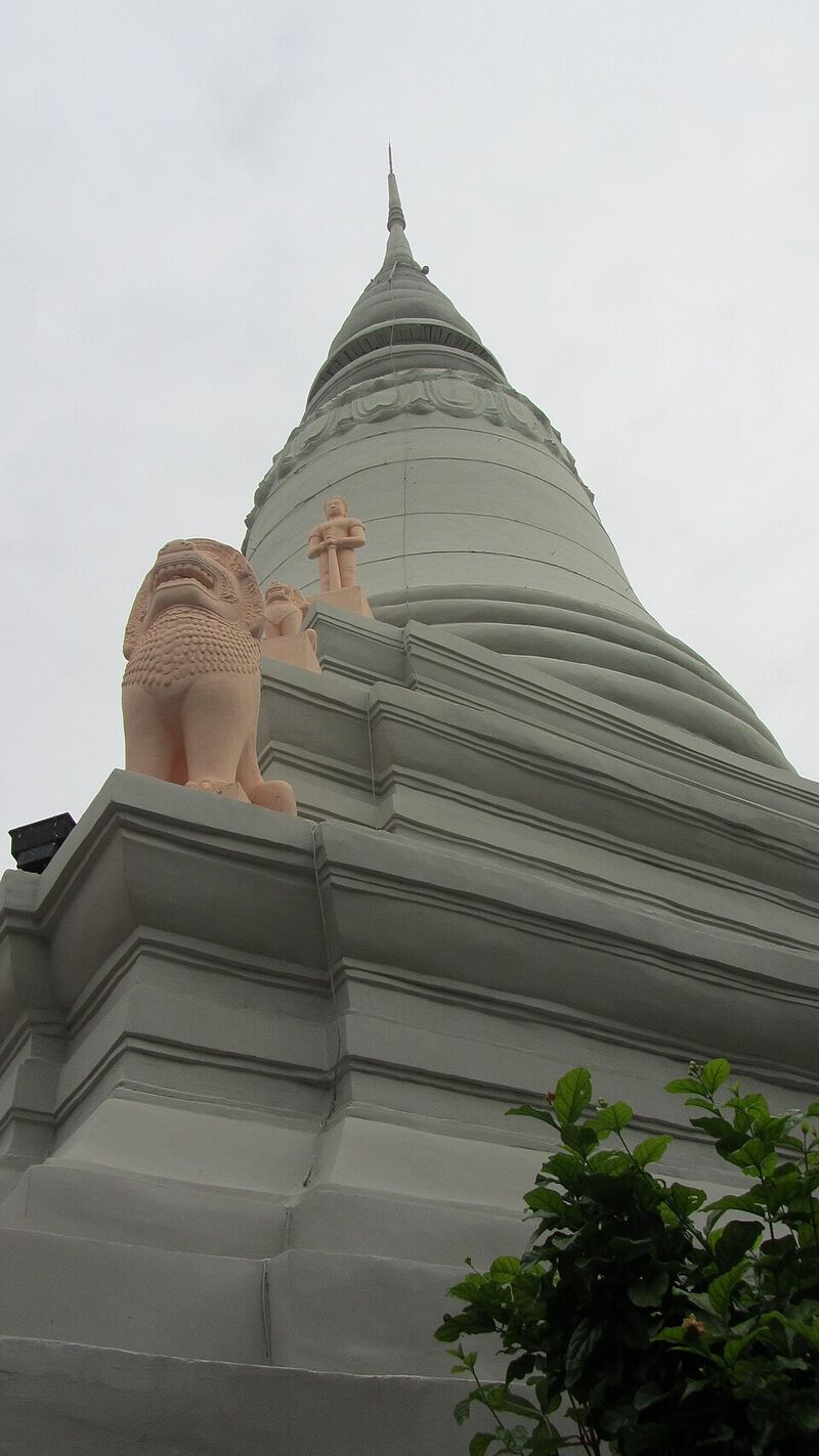 Low view of a pale grey-green bell-shaped stupa with a tall spire, with pink stone lions on the terrace at its base, under a pale grey sky.