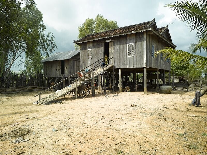 A weathered wooden house on stilts with a steep roof and a wooden ramp leading to the raised front door