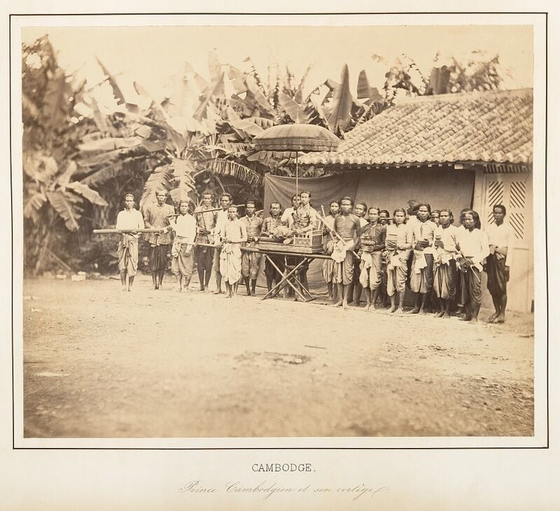 A prince seated on a carrying chair under a tall parasol, with a row of barefoot attendants in sampot on both sides holding poles, boxes and cups, in front of a tiled house and banana trees.