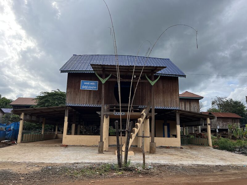 A two-storey wooden hall on stilts with a blue metal roof and a small signboard, standing on bare ground under a cloudy sky.