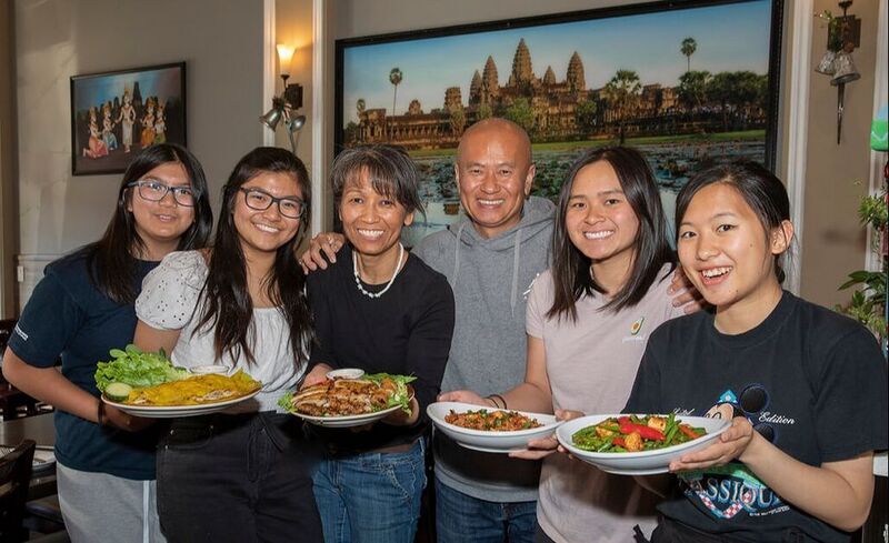 Six people smiling in a restaurant dining room, holding plates of food, in front of a large photo of Angkor Wat.