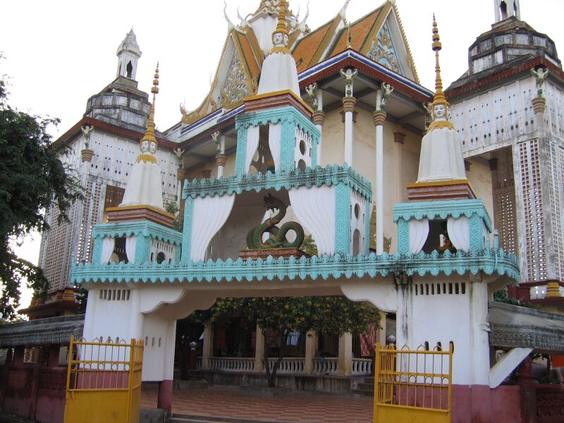 A white Khmer temple hall with teal trim and gilded roof spires behind small turquoise stupas and a yellow gate.
