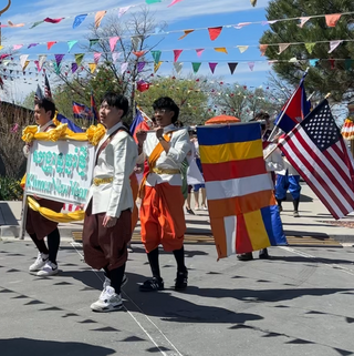 People in traditional dress walk in a procession carrying a sign and Buddhist, Cambodian and US flags.