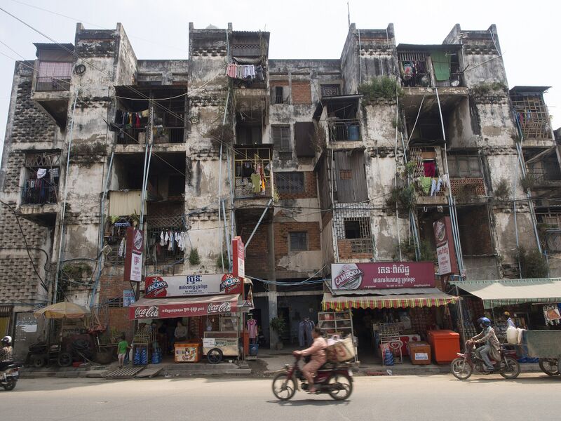 A weathered concrete apartment block with balconies and shop signs at street level, and motorbikes passing.