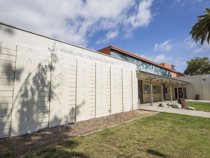 A white patterned block wall lettered Homeland Cultural Center beside a covered entrance under a blue sky with a palm tree.