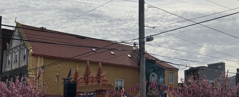 A yellow temple building with a red tile roof, a carved Khmer-style gate with three spires, a mural of the Buddha and pink blossoming trees under a cloudy sky.