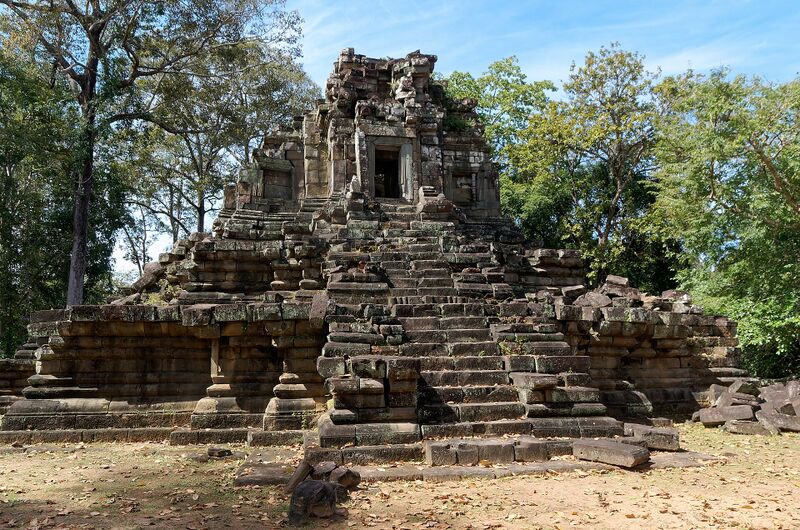 A ruined sandstone temple tower on a stepped terrace with a steep central stairway, among trees under a blue sky.