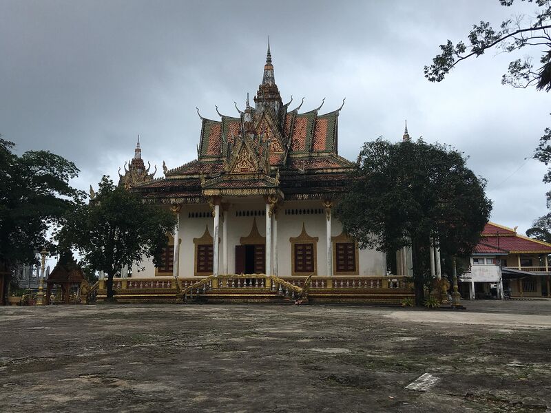 A white temple hall with gold trim and tiered pointed roofs behind a paved yard under a cloudy sky.
