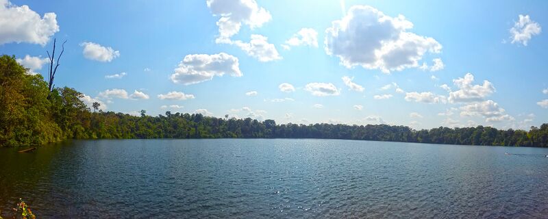 A wide round lake ringed by forest under a blue sky with white clouds.