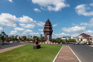 A tall tiered reddish-brown tower in the style of an Angkorian temple stands on a traffic circle with a few cars and motorbikes around it, seen across a grassy median with a stone lion statue, under a blue sky with white clouds.