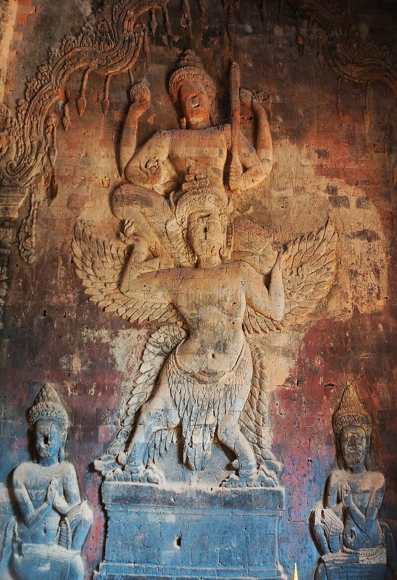 A large figure with several arms carved into a reddish brick wall, standing on the shoulders of a bird-like figure, with two smaller kneeling figures below.