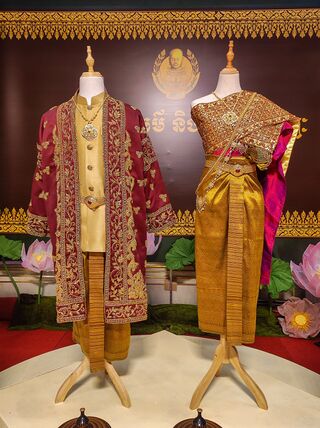 Two mannequins in front of a backdrop, one in a red and gold jacket, the other in a gold sampot and shawl.
