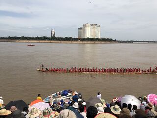 A crowd with umbrellas on a riverbank watching a long boat pass.