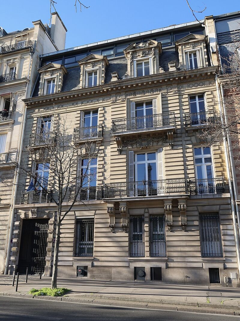 A six-storey cream stone apartment building with wrought-iron balconies, on a tree-lined Paris street.