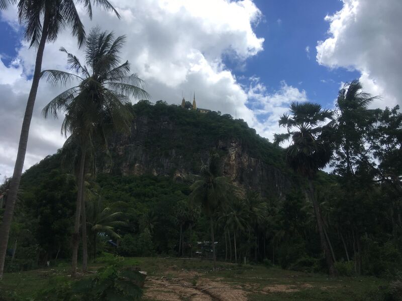 A steep wooded hill with a bare rock face rises behind tall palm trees and a dirt clearing. Two golden spires and a building stand on the summit against a sky of white clouds.