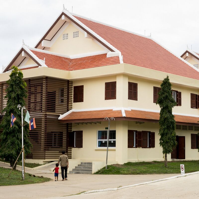 A two-storey cream building with red tiled roofs and Lao flags, a man and small child walking toward it.