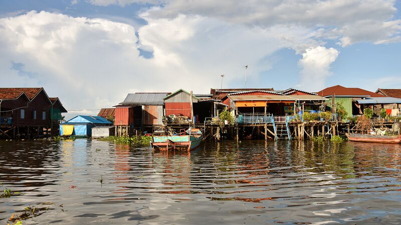 A row of wooden houses with tin roofs raised on stilts above brown floodwater, under a sky of large white clouds.