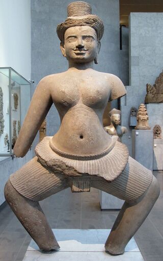 A sandstone statue of a female figure in a dance pose, arms missing, on a museum plinth.