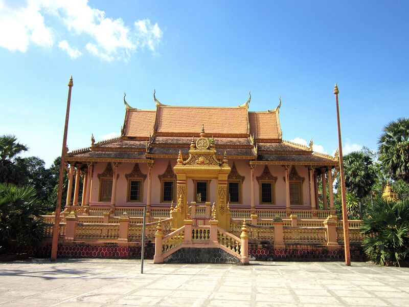 A salmon-coloured Khmer temple hall with a tiered roof, gilded entrance stairway and two tall flagpoles under a blue sky.