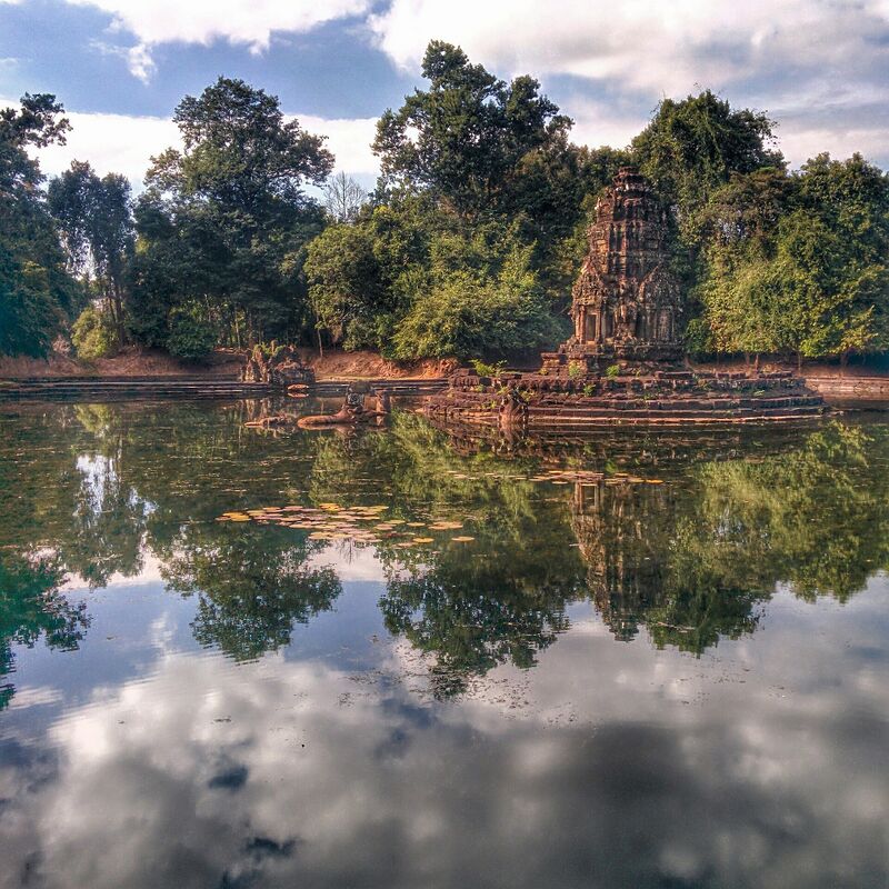 A small stone tower on a low island in a still pool, with trees behind it and clouds reflected in the water.