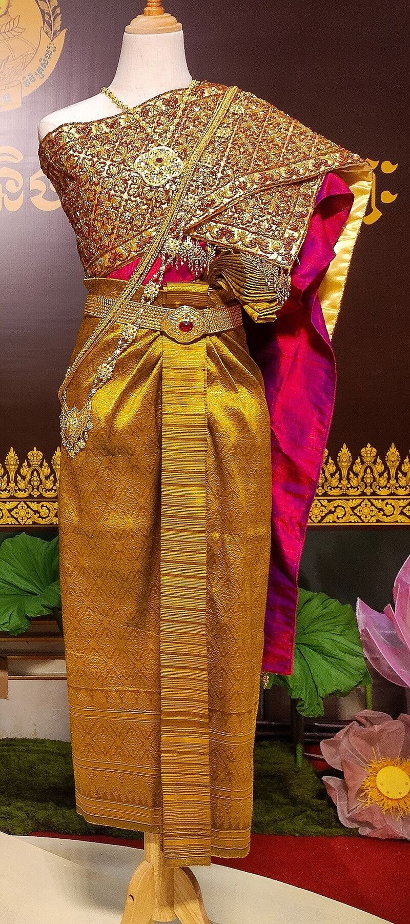 A gold brocade sbai draped over one shoulder of a dress stand, above a gold sampot and a pink sash.